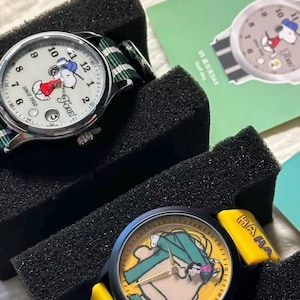 May include: Two watches featuring Snoopy designs. One watch has a white face with Snoopy in a red outfit, a green and white striped band, and the text "FORE!" and "SINCE 1950". The other watch has a yellow face with Snoopy in a green outfit and the text "HAAA".