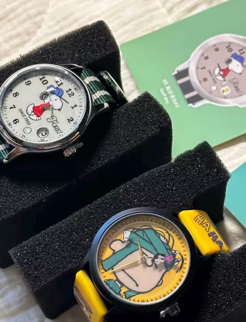 May include: Two watches featuring Snoopy designs. One watch has a white face with Snoopy in a red outfit, a green and white striped band, and the text "FORE!" and "SINCE 1950". The other watch has a yellow face with Snoopy in a green outfit and the text "HAAA".