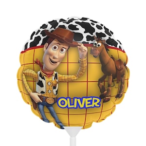 K&ouml;nnte beinhalten: Ein runder, gelber Ballon mit der Figur Woody aus Toy Story, mit braunem Cowboyhut und Weste. Der Ballon hat einen schwarz-wei&szlig;en Kuhdruckrand und den Namen "OLIVER" in blauen Buchstaben.