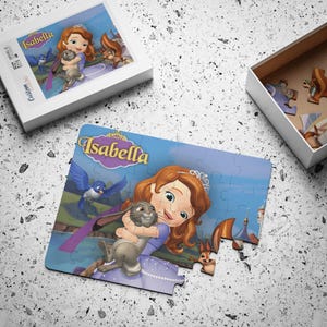 May include: A partially assembled jigsaw puzzle featuring a cartoon princess, a rabbit, and a squirrel, with the name "Isabella" in a decorative font. The puzzle is on a light gray surface, with the puzzle box and a box of puzzle pieces visible.
