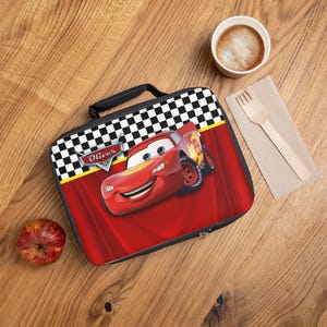 Puede incluir: Una bolsa de almuerzo roja y negra con Rayo McQueen de la pel&iacute;cula Cars. La bolsa tiene un asa negra, un dise&ntilde;o de bandera a cuadros y el nombre "Oliver". Una manzana, una taza de caf&eacute; y un tenedor est&aacute;n sobre la superficie de madera.