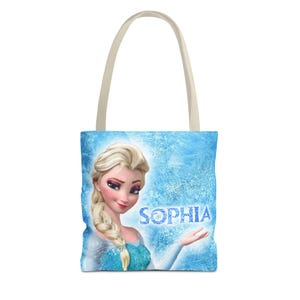 以下が含まれることがあります： ブロンドの女性の肖像画と「SOPHIA」という名前が青色で描かれた、水色のトートバッグ。バッグには薄いベージュのハンドルと、霜で覆われた氷のデザインの青い背景があります。