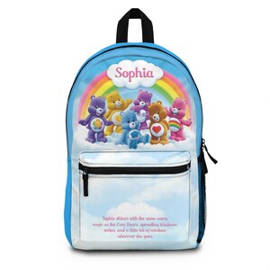May include: A blue backpack featuring a rainbow and Care Bears graphic. The backpack has a black handle and a front pocket with a zipper. The name "Sophia" is written above the Care Bears.