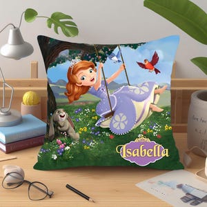May include: A decorative pillow featuring a cartoon illustration of a girl on a swing in a garden. The girl wears a purple dress and has red hair. The pillow has the name "Isabella" on it.