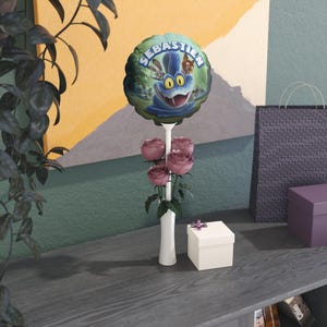 以下が含まれることがあります： 「SEBASTIEN」という名前と漫画のキャラクターが描かれた風船と、白い花瓶に入ったピンクのバラの装飾。リボン付きの小さなギフトボックスが近くにあります。