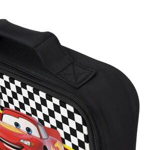 Puede incluir: Bolsa de almuerzo negra con un dise&ntilde;o de coche de carreras rojo y un patr&oacute;n de bandera a cuadros. Tiene un asa negra y un cierre de cremallera. El coche de carreras tiene ojos azules y un dise&ntilde;o amarillo y rojo.