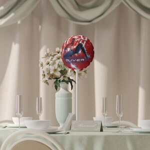 May include: A festive table setting with a red balloon featuring a superhero graphic and the name "OLIVER". The centerpiece includes white flowers in a light green vase, champagne flutes, and a "Reserved" sign.