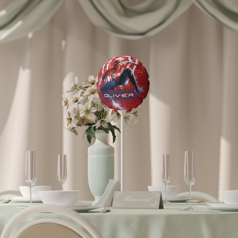 May include: A festive table setting with a red balloon featuring a superhero graphic and the name "OLIVER". The centerpiece includes white flowers in a light green vase, champagne flutes, and a "Reserved" sign.