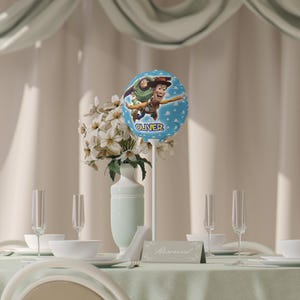 K&ouml;nnte beinhalten: Eine Tischdekoration mit einem hellblauen Ballon mit Woody und Buzz Lightyear aus Toy Story, mit dem Namen "Oliver". Der Ballon ist Teil eines Blumenschmucks mit wei&szlig;en Blumen in einer hellblauen Vase. Sektgl&auml;ser und Gedecke sind sichtbar.