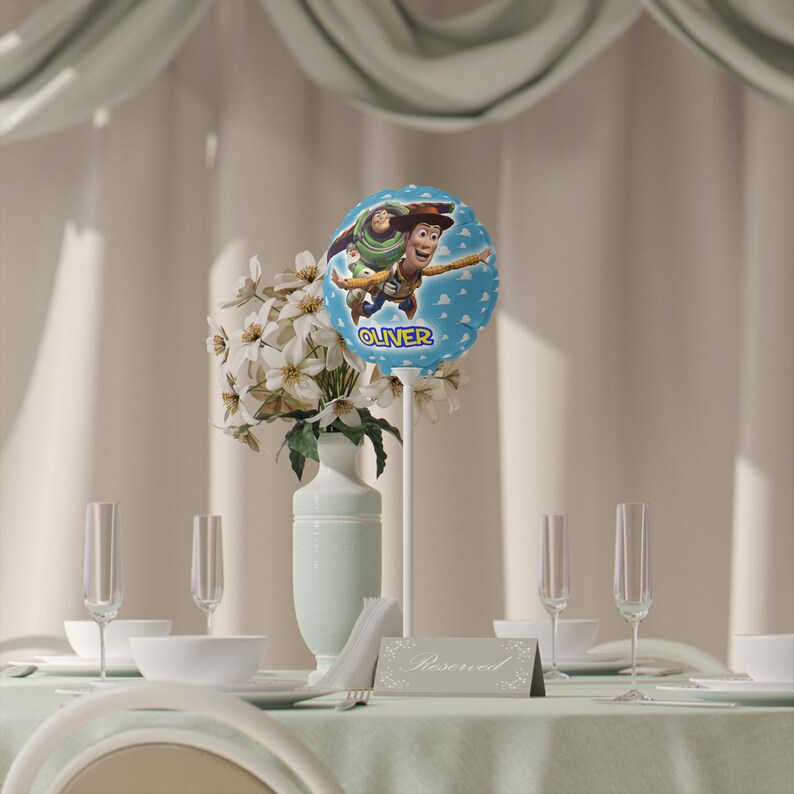 K&ouml;nnte beinhalten: Eine Tischdekoration mit einem hellblauen Ballon mit Woody und Buzz Lightyear aus Toy Story, mit dem Namen "Oliver". Der Ballon ist Teil eines Blumenschmucks mit wei&szlig;en Blumen in einer hellblauen Vase. Sektgl&auml;ser und Gedecke sind sichtbar.