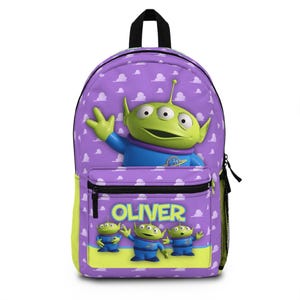 May include: Purple backpack featuring a green alien character with three eyes and a blue outfit. The backpack has a front pocket with the name "OLIVER" in yellow letters. The background is a pattern of white clouds.