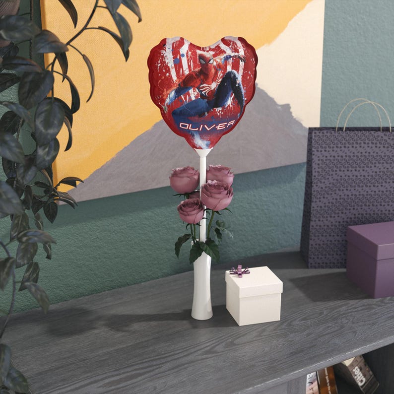 May include: A heart-shaped red balloon featuring Spider-Man, with the name "OLIVER" printed on it, is held above a bouquet of pink roses in a white vase. A small gift box with a purple ribbon sits nearby.