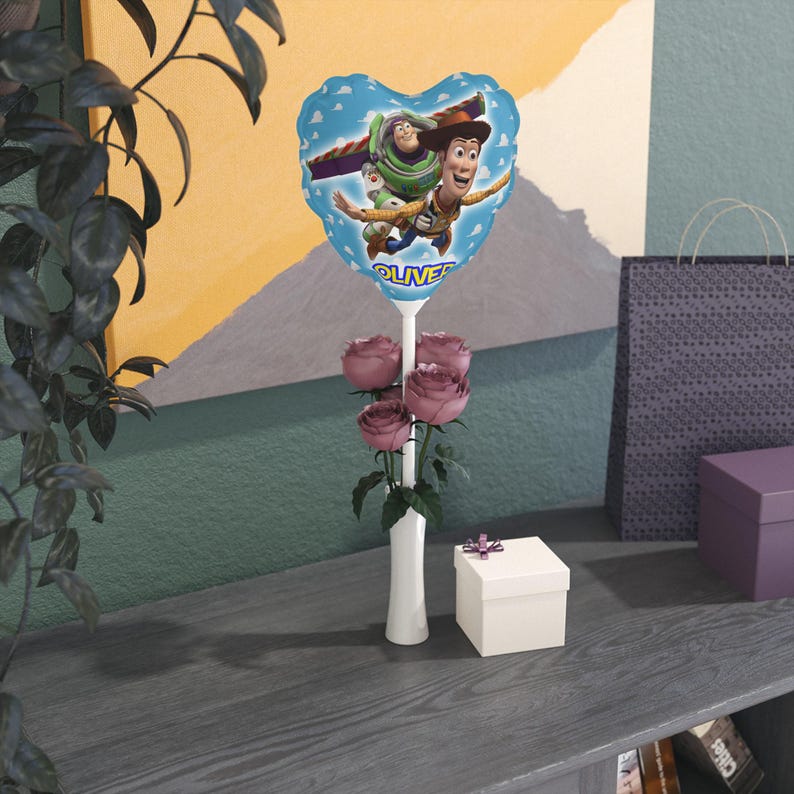 K&ouml;nnte beinhalten: Ein herzf&ouml;rmiger blauer Ballon mit Buzz Lightyear und Woody aus Toy Story, mit dem Namen "OLIVER". Der Ballon ist Teil eines Blumenarrangements mit rosa Rosen in einer wei&szlig;en Vase und einer kleinen Geschenkbox.