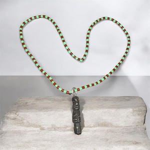 Puede incluir: Un collar con una larga hilera de cuentas cilíndricas que alternan el verde menta y el rojo terracota. Un colgante gris oscuro, tallado en forma de tótem vertical, cuelga de una anilla de metal plateado en el centro de la hilera.