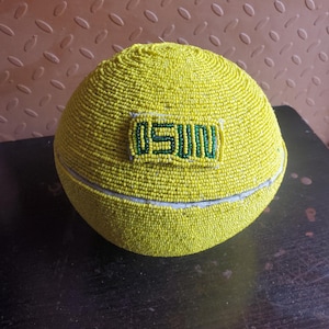 May include: A yellow beaded hat with the word "OSUN" in green on the front. The hat is round and covered in small yellow beads. The hat is sitting on a dark surface.