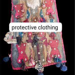 Pode incluir: Vestuário de proteção feito à mão com o texto "protective clothing". O tecido apresenta detalhes em vermelho, azul e branco, elementos decorativos e enfeites de conchas. A peça tem uma borda rosa e pompons azuis pendurados.