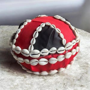 Puede incluir: Un sombrero de tela roja y negra adornado con hileras de conchas blancas. El sombrero tiene una forma circular con un patrón segmentado de tela roja y negra, y las conchas están cosidas a lo largo de los bordes.
