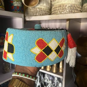 May include: A light blue beaded cap with geometric patterns in red, black, white, and yellow. The cap has a small beaded ball on top and a red tassel with white fringe. This traditional headwear is likely from an African culture.