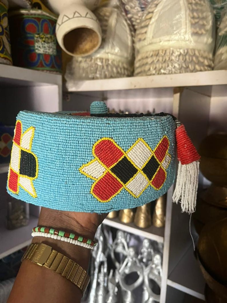 May include: A light blue beaded cap with geometric patterns in red, black, white, and yellow. The cap has a small beaded ball on top and a red tassel with white fringe. This traditional headwear is likely from an African culture.