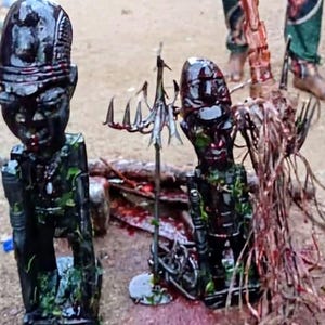 May include: Two dark wooden statues with detailed carvings, possibly representing figures. One statue is on the left, and another is on the right. A metal object with spikes is between them. The statues are surrounded by red and brown organic material.