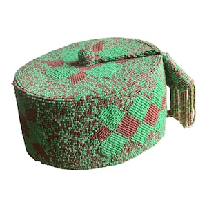 May include: An oval-shaped hat, beaded in green and maroon. The hat has a geometric pattern and a small, beaded handle with fringe. The hat is likely handmade, with a unique, textured appearance.