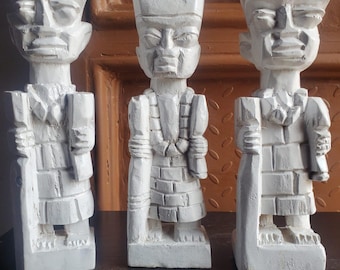May include: Three white, carved wooden statues of figures wearing crowns and ornate clothing. The statues have a weathered appearance and stand on a dark surface. The figures have a regal appearance, with detailed facial features and attire.