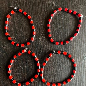 May include: Four circular bracelets arranged in a two-by-two grid. Each bracelet is constructed from alternating cylindrical red and black beads strung on a white cord. The beads are uniform in size and shape, creating a repeating color pattern.