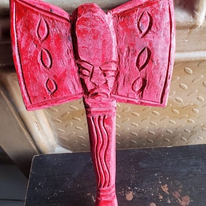 May include: A carved wooden axe painted red. The axe head features a stylized face and two wing-like extensions with carved oval shapes. The handle is carved with a wavy design and a rounded base. The axe is displayed on a dark surface.