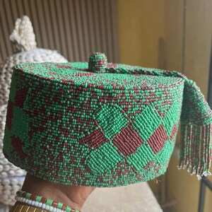 May include: A green and maroon beaded hat with a geometric design. The hat is cylindrical with a small, round top and a fringed edge. The hat is held in a hand, with gold and beaded bracelets visible. The hat is made of small beads.
