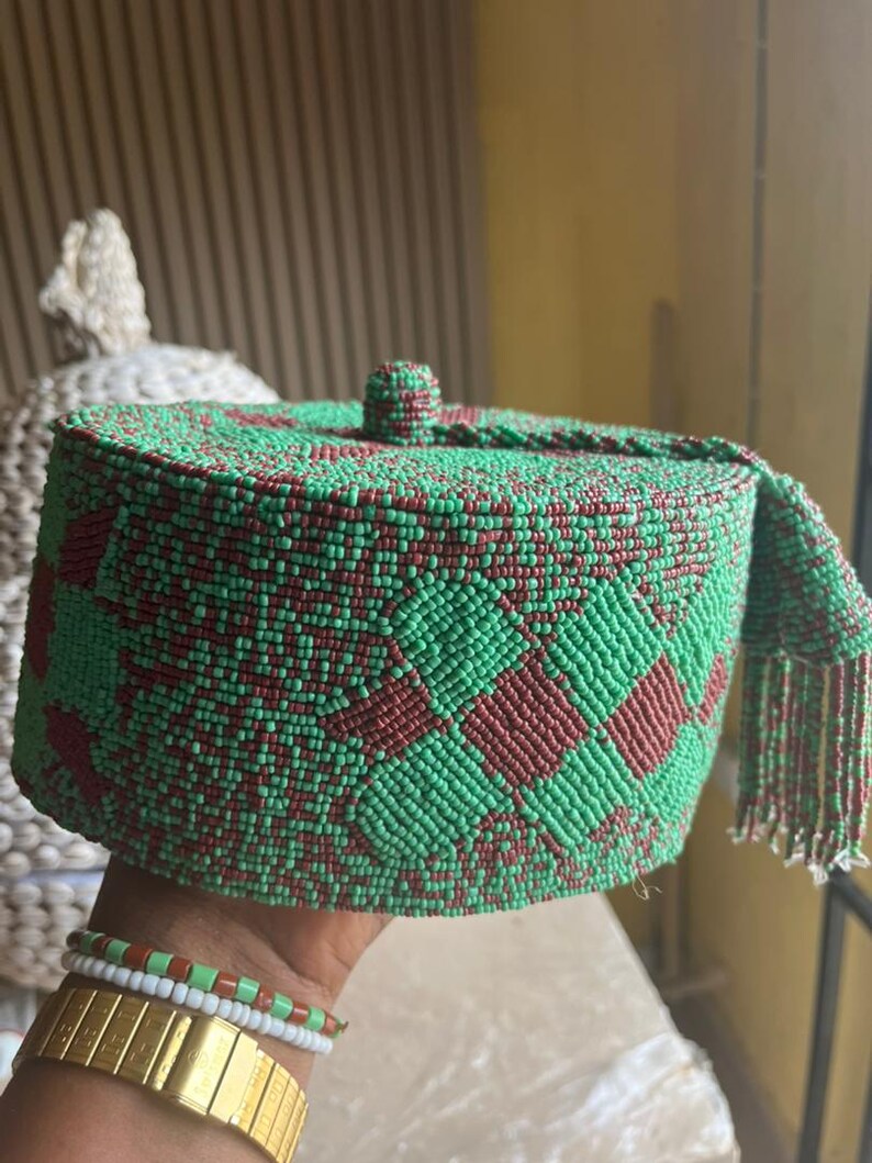 May include: A green and maroon beaded hat with a geometric design. The hat is cylindrical with a small, round top and a fringed edge. The hat is held in a hand, with gold and beaded bracelets visible. The hat is made of small beads.