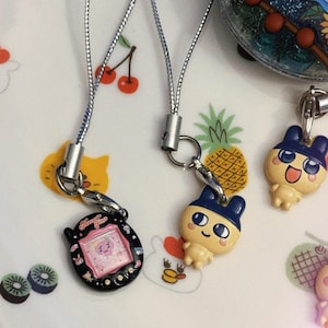 May include: A collection of colorful, small, vintage handheld game charms with silver cords. One charm is black with a pink screen, another is a yellow cartoon character with a blue hat. The background is a white plate with fruit and animal illustrations.