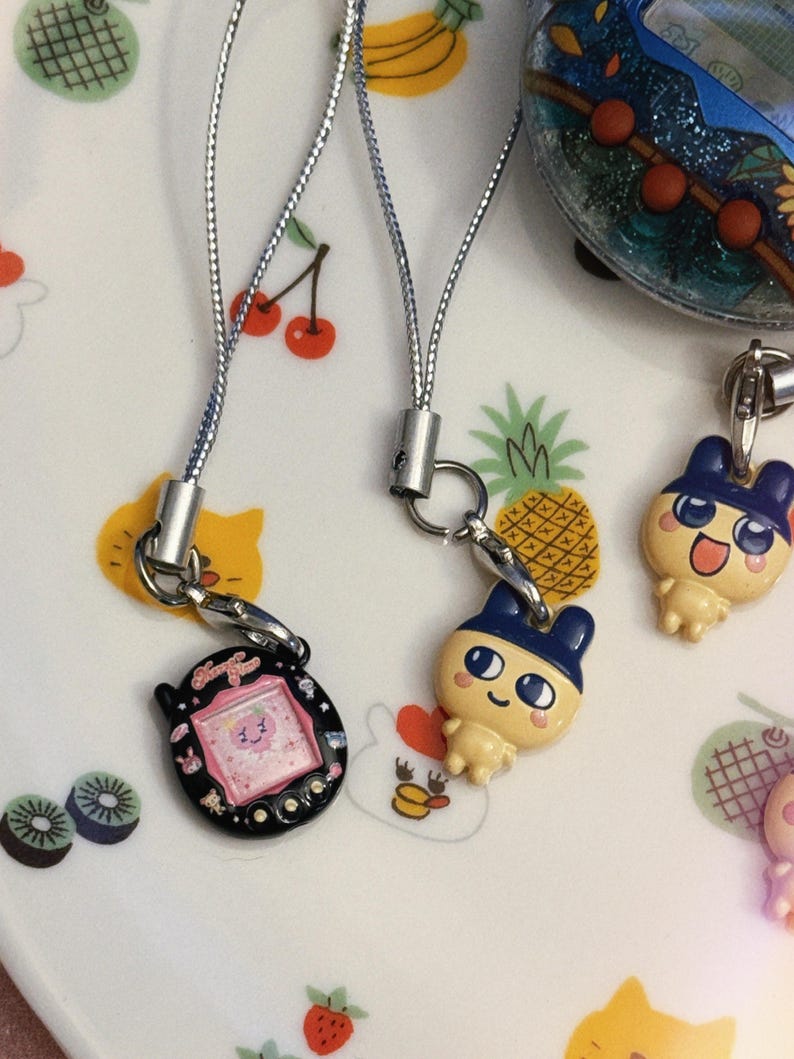 May include: A collection of colorful, small, vintage handheld game charms with silver cords. One charm is black with a pink screen, another is a yellow cartoon character with a blue hat. The background is a white plate with fruit and animal illustrations.