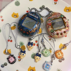 May include: A collection of Tamagotchi virtual pet devices and charms. The devices feature screens displaying pixelated characters. The charms are in various colors and designs, including cartoon characters. The items are on a white plate with fruit and cat illustrations.