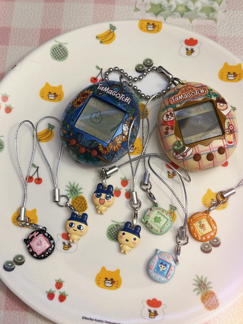 May include: A collection of Tamagotchi virtual pet devices and charms. The devices feature screens displaying pixelated characters. The charms are in various colors and designs, including cartoon characters. The items are on a white plate with fruit and cat illustrations.
