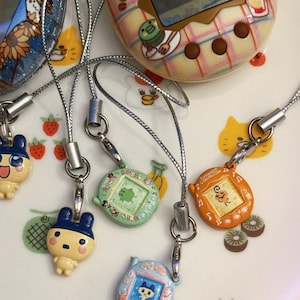 May include: A collection of vintage handheld digital pet game devices in various colors, including blue, green, and orange. Each device has a small screen and is attached to a silver lanyard. The background features cartoon illustrations.