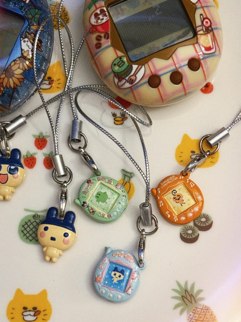 May include: A collection of vintage handheld digital pet game devices in various colors, including blue, green, and orange. Each device has a small screen and is attached to a silver lanyard. The background features cartoon illustrations.