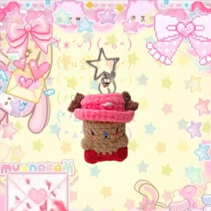 May include: A handmade crochet keychain featuring a brown bear-like character wearing a pink hat and red shoes. The keychain has a silver star-shaped clasp. The design is whimsical and cute, perfect for adding a touch of charm to keys or bags.