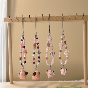 May include: Four beaded phone charms with pink, white, and brown beads, and small charms. Each charm has a different pink cartoon character at the bottom. The charms hang from a wooden display stand.