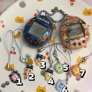 May include: A collection of Tamagotchi virtual pet devices and charms. Two larger devices with screens are connected to smaller charms numbered 1-7. The devices and charms feature various colors and designs, including floral patterns and cartoon characters.