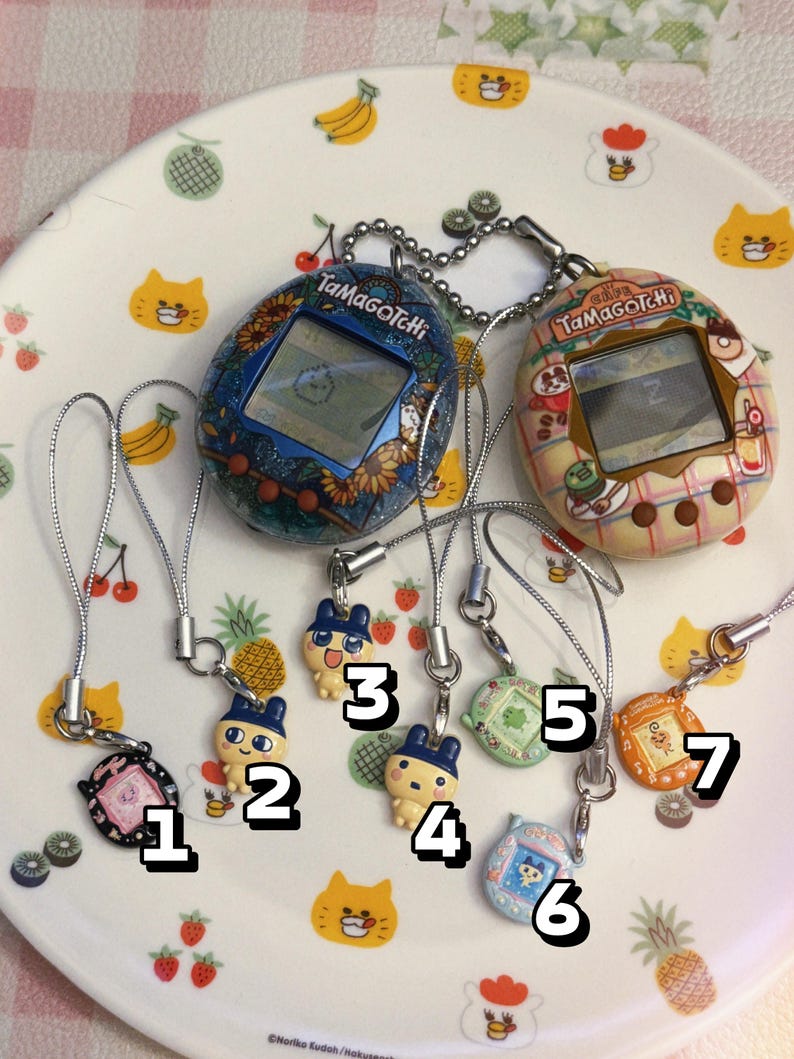 May include: A collection of Tamagotchi virtual pet devices and charms. Two larger devices with screens are connected to smaller charms numbered 1-7. The devices and charms feature various colors and designs, including floral patterns and cartoon characters.