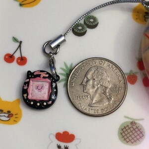 May include: A black and pink handheld electronic game charm with a silver chain next to a US quarter. The game has a small screen with a cartoon character. The background is a white plate with fruit illustrations.