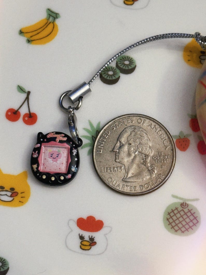 May include: A black and pink handheld electronic game charm with a silver chain next to a US quarter. The game has a small screen with a cartoon character. The background is a white plate with fruit illustrations.