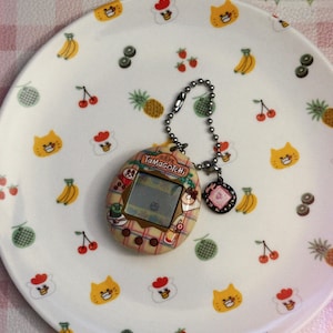 May include: A vintage Tamagotchi digital pet with a silver chain and charm rests on a white plate. The plate is decorated with colorful cartoon fruits and cat faces. The Tamagotchi has a small screen and a patterned design.