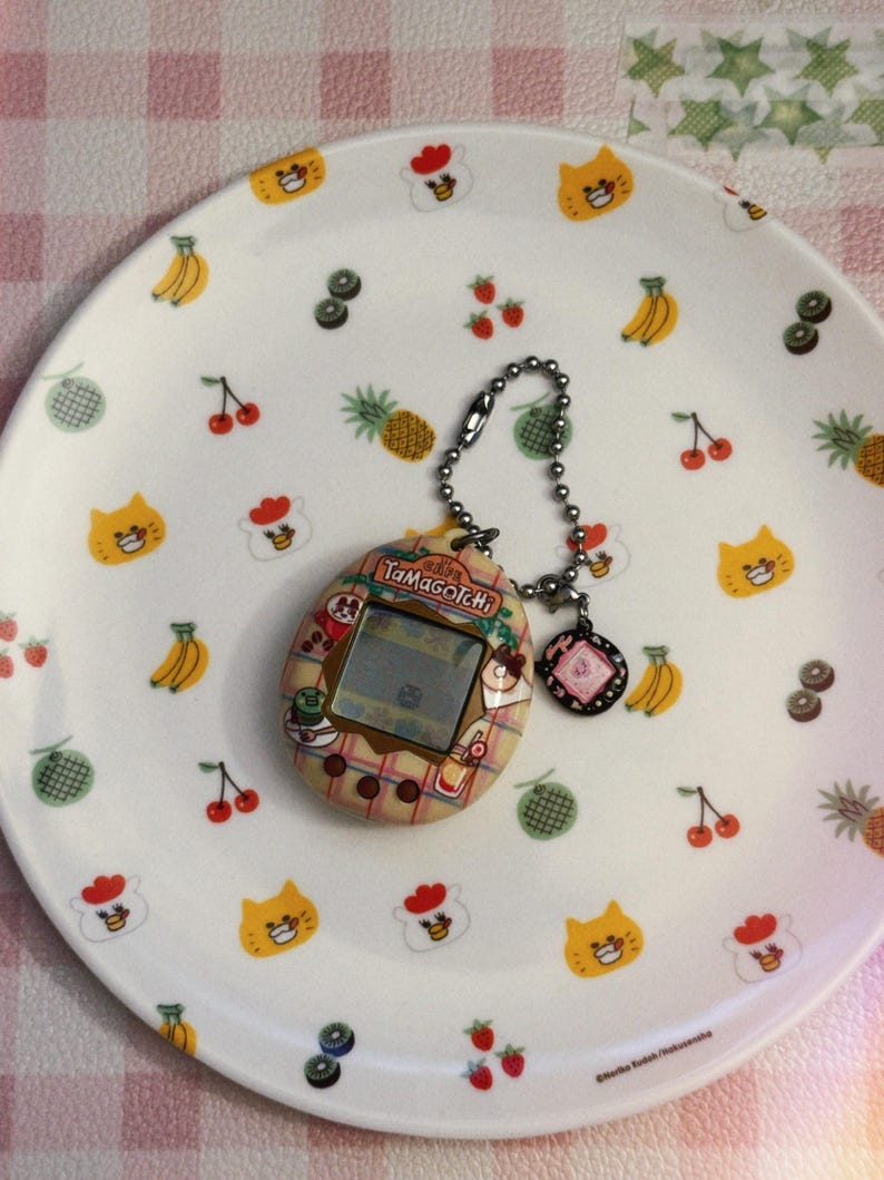 May include: A vintage Tamagotchi digital pet with a silver chain and charm rests on a white plate. The plate is decorated with colorful cartoon fruits and cat faces. The Tamagotchi has a small screen and a patterned design.