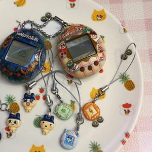 May include: Two vintage Tamagotchi digital pets, one blue with floral design, the other beige with a patterned design, sit on a decorative plate. Several small character charms dangle from the devices, adding a playful touch to the collection.