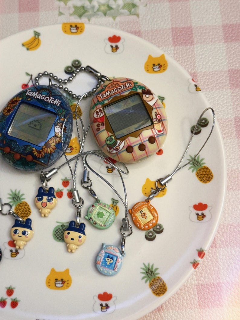 May include: Two vintage Tamagotchi digital pets, one blue with floral design, the other beige with a patterned design, sit on a decorative plate. Several small character charms dangle from the devices, adding a playful touch to the collection.