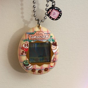 May include: A vintage Tamagotchi virtual pet device with a cafe theme. The egg-shaped device features a screen, buttons, and a decorative design with food-related graphics. It hangs from a silver chain with a charm.