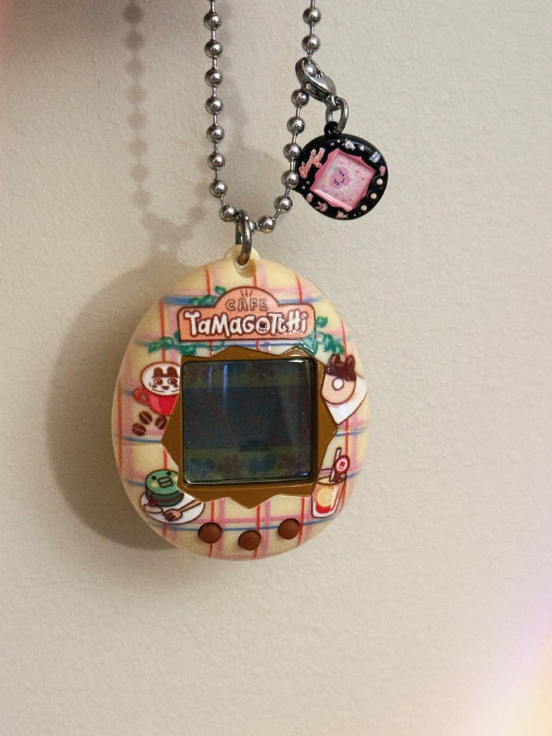 May include: A vintage Tamagotchi virtual pet device with a cafe theme. The egg-shaped device features a screen, buttons, and a decorative design with food-related graphics. It hangs from a silver chain with a charm.