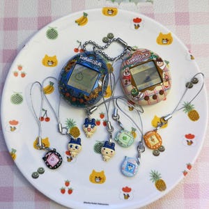 May include: A collection of Tamagotchi virtual pet devices and charms on a decorative plate. The devices feature screens and are in various colors, including blue, pink, and brown. The plate has a fruit and cat design.