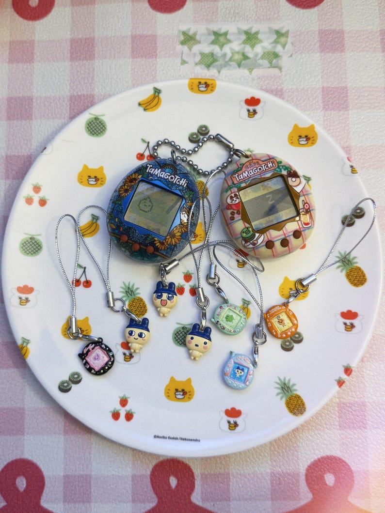 May include: A collection of Tamagotchi virtual pet devices and charms on a decorative plate. The devices feature screens and are in various colors, including blue, pink, and brown. The plate has a fruit and cat design.
